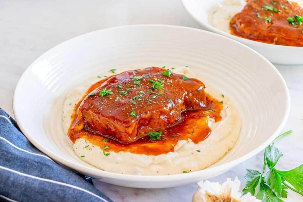 A bowl of mashed potatoes topped with a piece of pork covered in brown gravy, garnished with chopped herbs&mdash;perfect for pork dinner ideas.