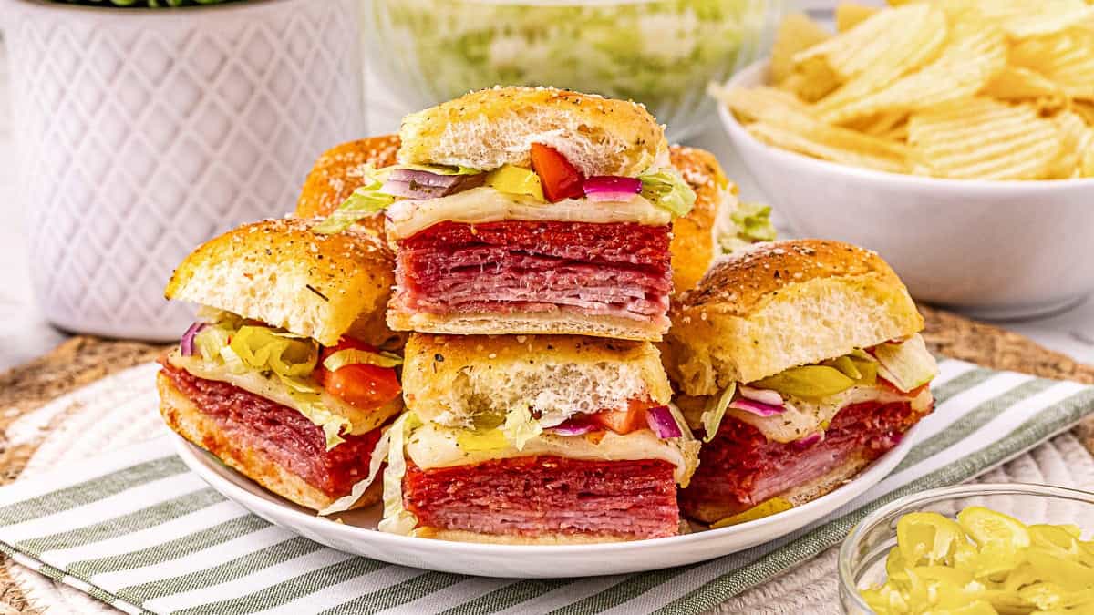A plate of stacked Italian sub sliders filled with deli meats, cheese, lettuce, tomatoes, and peppers, with a bowl of potato chips in the background.