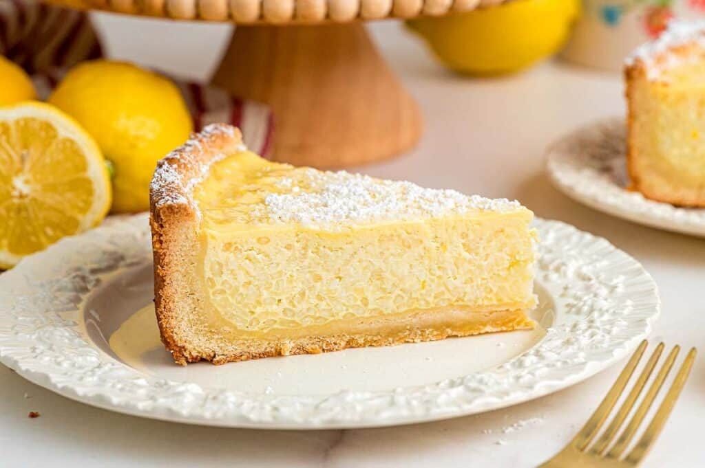 A slice of lemon ricotta pie with a dusting of powdered sugar sits on a white plate; this easy dessert is perfect for any occasion, with lemons and another slice in the background.