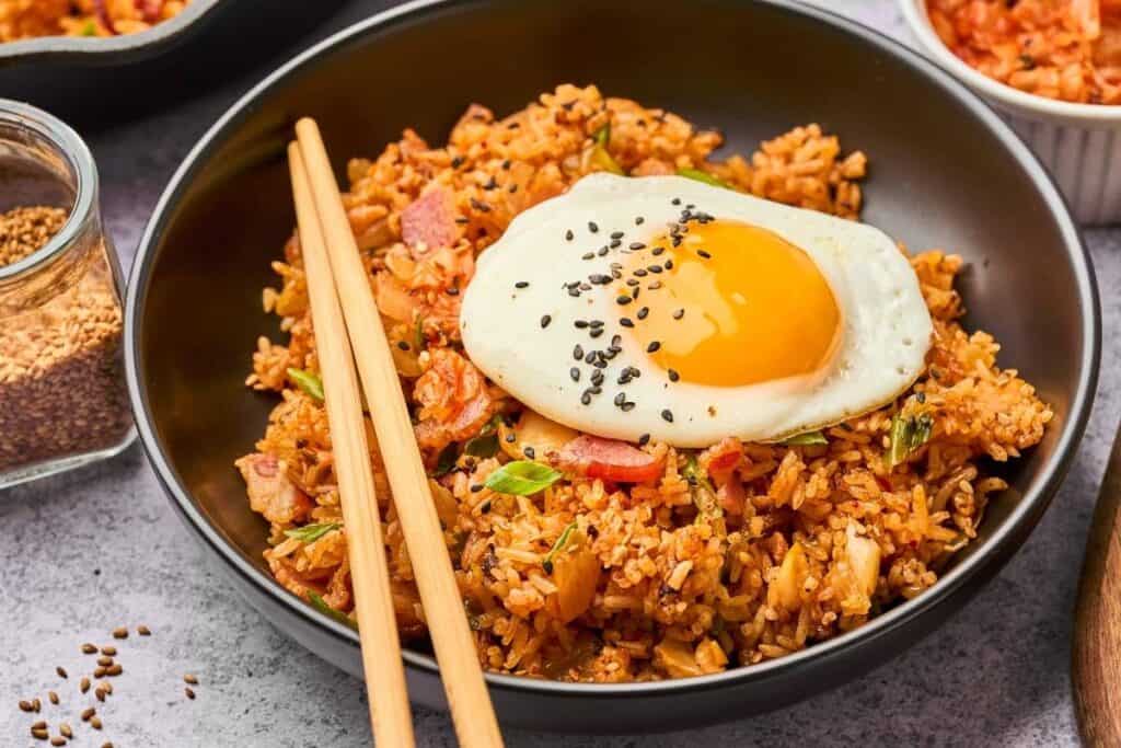 A bowl of kimchi fried rice topped with a sunny-side-up egg and black sesame seeds, served with chopsticks—perfect for low effort dinners.