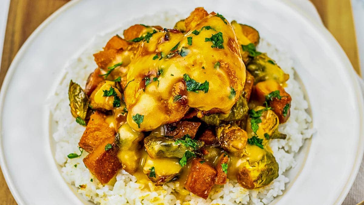 A plate of white rice topped with roasted vegetables and a piece of chicken, covered in a yellow sauce and garnished with chopped parsley.