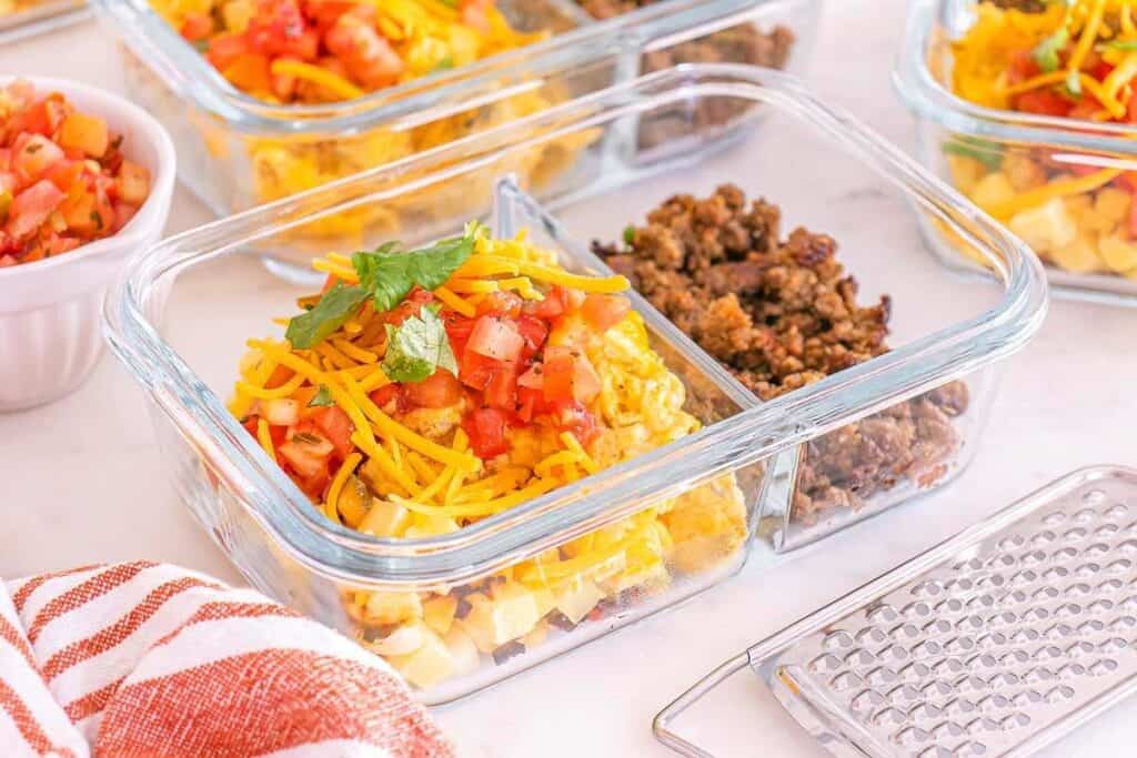 A glass meal prep container with shredded cheese, chopped tomatoes, and ground beef separated into compartments offers tasty breakfast ideas, alongside a small grater and a striped towel.