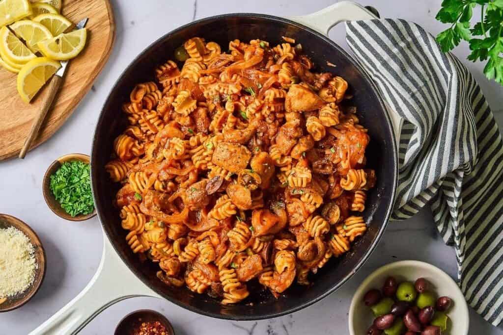A skillet filled with cooked rotini pasta, chicken pieces, olives, and tomato sauce—this Pasta Classics dish is surrounded by small bowls of olives, parmesan, herbs, lemon slices, and a striped towel.