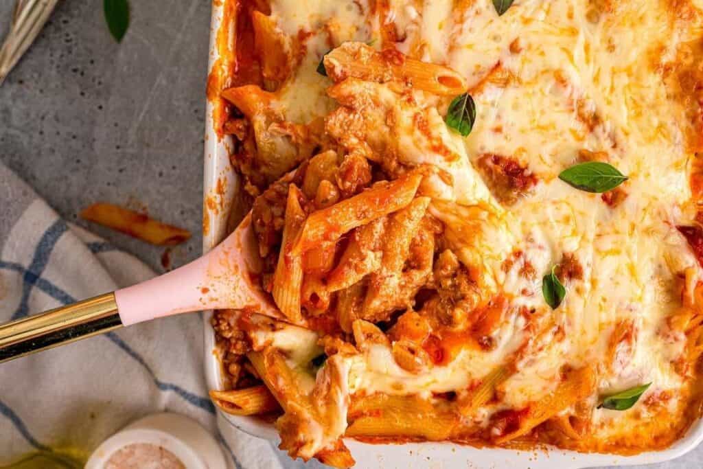 A Pasta Classics favorite: a baking dish of cheesy baked penne pasta with tomato sauce and ground meat, partially served with a pink-handled spoon.