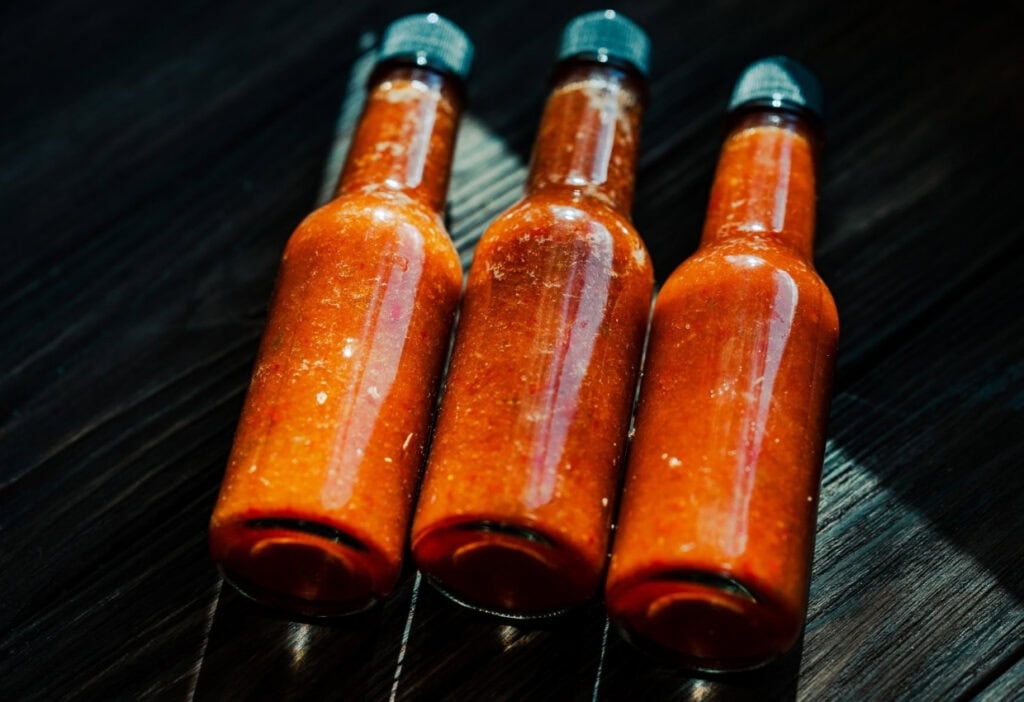 Three glass bottles filled with red hot sauce are arranged side by side on a dark wooden surface.