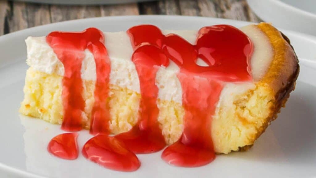 A slice of cheesecake topped with a thick layer of white cream and drizzled with bright red strawberry sauce on a white plate.