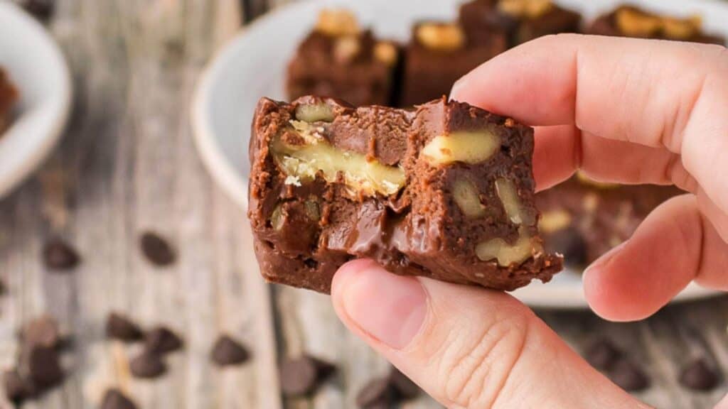 A hand holds a piece of chocolate fudge with visible walnuts; more fudge pieces are on a plate in the background.