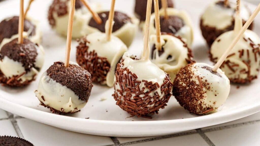A plate of chocolate cake pops, partially coated in white chocolate and decorated with chocolate sprinkles, each with a wooden stick inserted.
