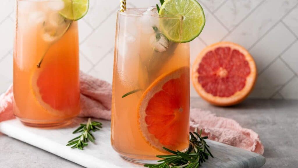 Two tall glasses of grapefruit and lime cocktail with ice, garnished with rosemary and citrus slices, sit on a marble tray with a halved grapefruit and rosemary sprigs nearby.