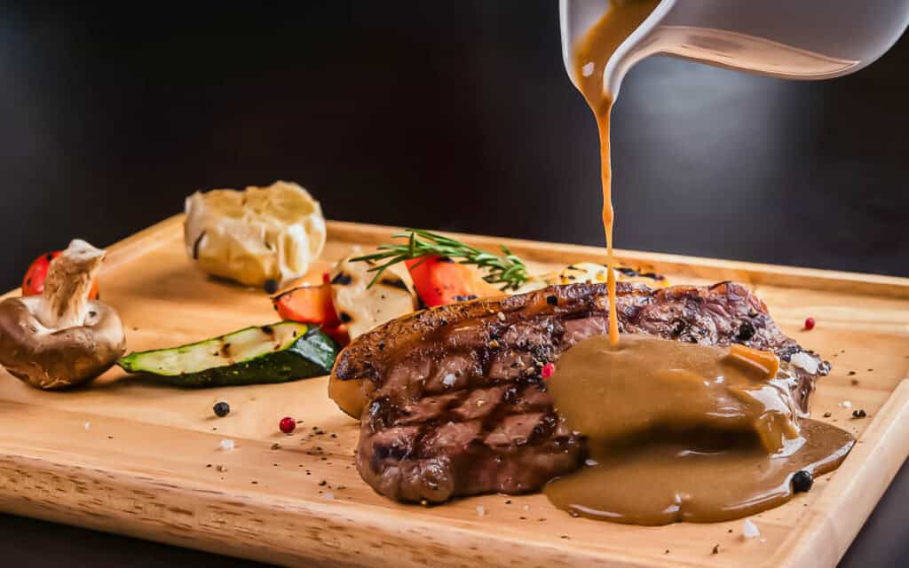 Gravy is poured over a grilled steak served on a wooden board with grilled vegetables, mushrooms, and garnish.