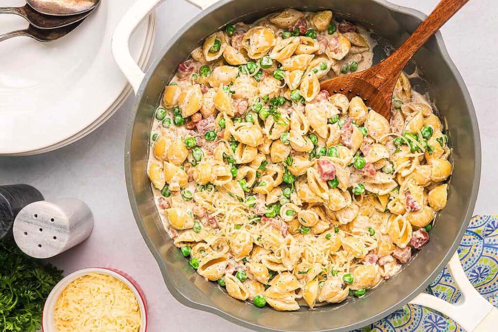 A pot of creamy shell pasta with peas and diced ham, perfect for a cozy pasta dinner, sits on a table with a wooden spoon, a bowl of grated cheese, and plates nearby.