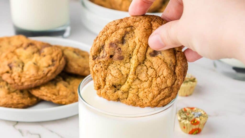 A hand dips a chocolate chip cookie into a glass of milk, with more cookies and mini peanut butter cups on a plate in the background.