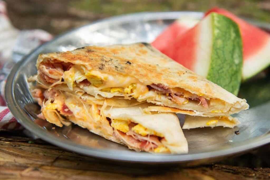 Two breakfast quesadilla wedges with eggs, ham, and cheese on a metal plate, served alongside a slice of watermelon&mdash;a delicious option for anyone searching for tasty breakfast ideas.