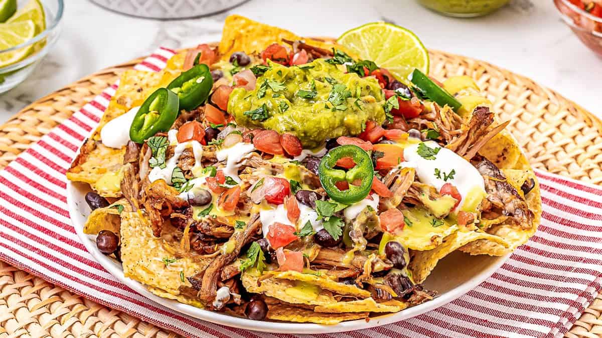 A plate of nachos topped with shredded meat, black beans, diced tomatoes, jalape&ntilde;os, guacamole, sour cream, and chopped cilantro, served on a red striped cloth.