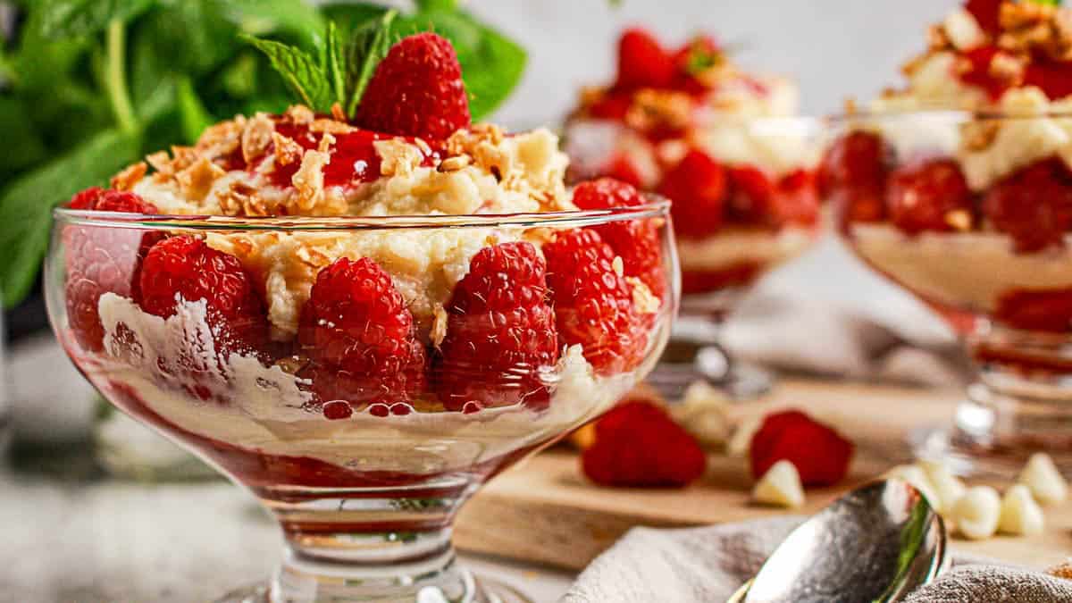 Glass dessert bowls filled with layers of fresh raspberries, creamy mixture, and granola, garnished with mint leaves and extra raspberries, sit on a table beside a spoon.