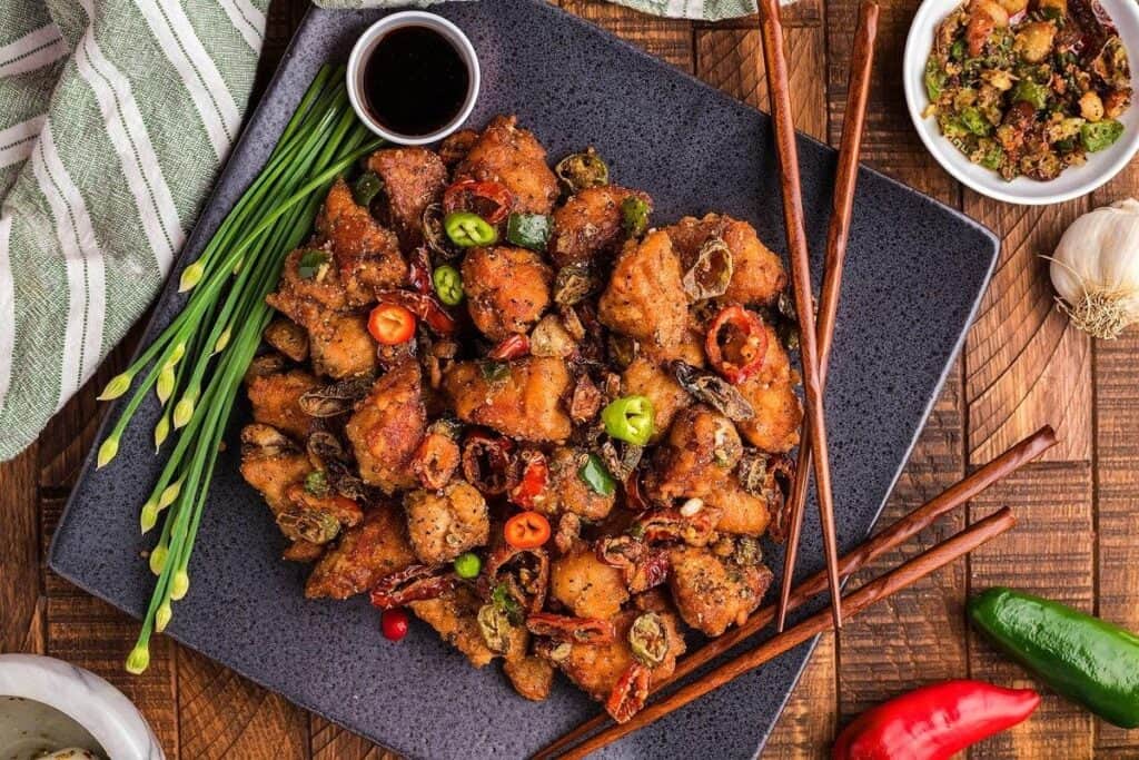 A square black plate of fried chicken pieces garnished with sliced green onions and red chilies, served with dipping sauce and wooden chopsticks on a wooden table&mdash;perfect inspiration alongside your favorite Pork Dinner ideas.