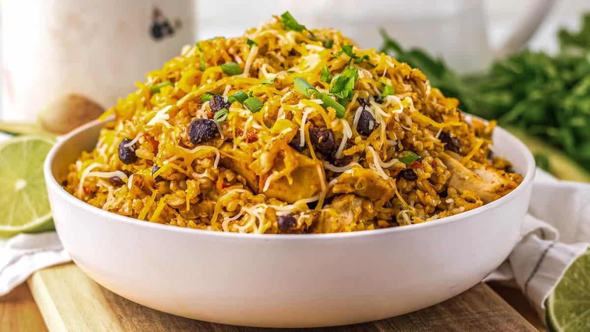 A bowl of rice mixed with black beans, shredded chicken, vegetables, spices, and garnished with chopped herbs and shredded cheese.