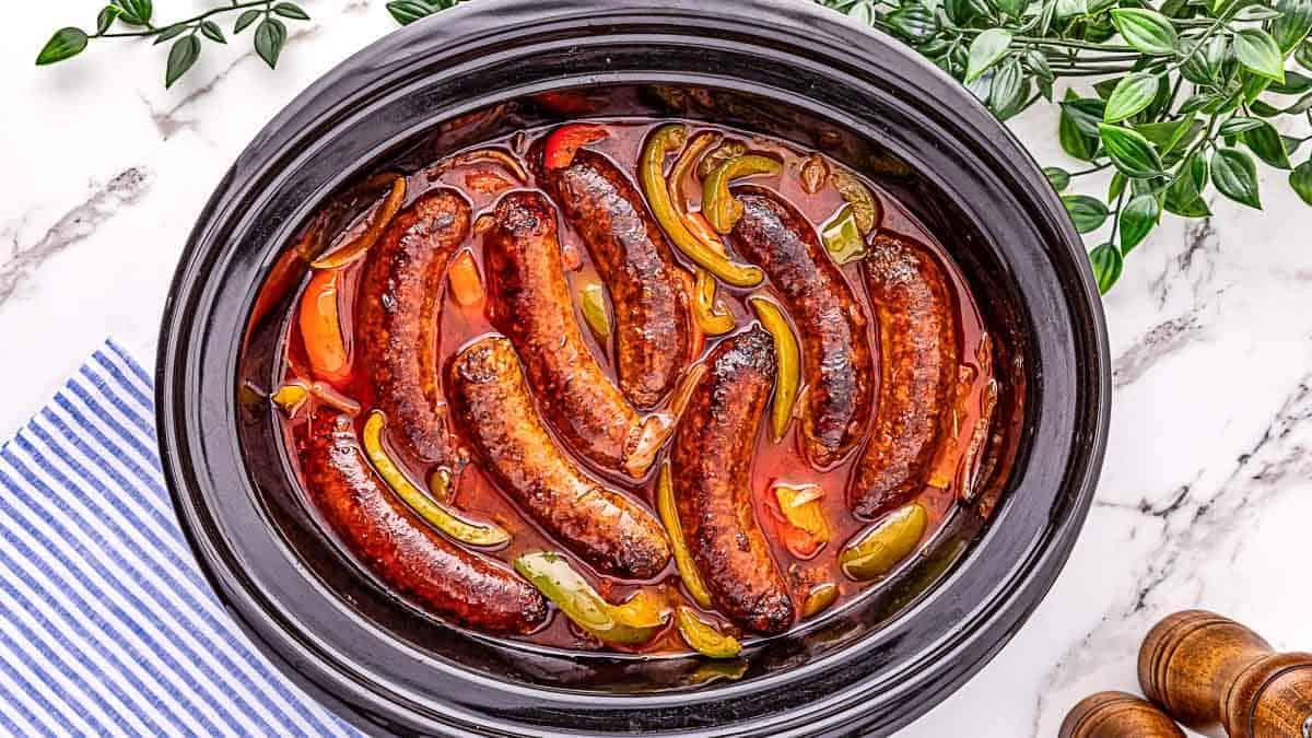 Slow cooker with cooked sausages, sliced bell peppers, and tomato sauce on a marble surface, with green plants and a striped cloth nearby.