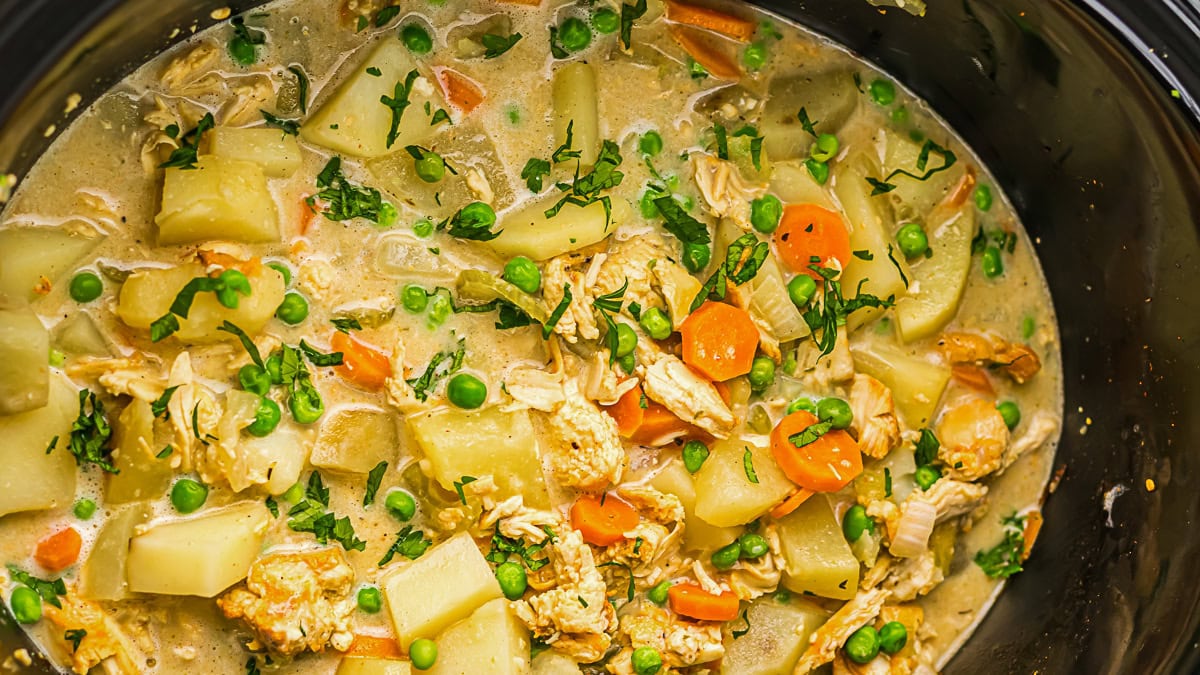 A close-up of a slow cooker filled with a creamy stew containing chunks of potatoes, carrots, peas, shredded chicken, and garnished with fresh herbs.