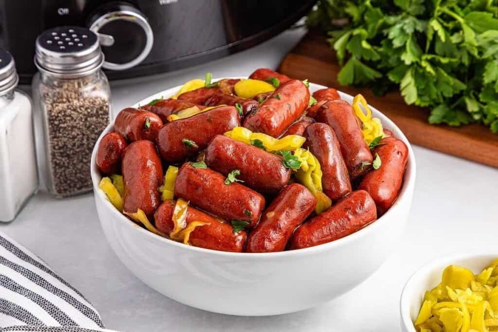 A white bowl filled with cooked cocktail sausages and sliced pepperoncini, perfect for Game Day Snacks, sits on a countertop next to salt, pepper, and fresh parsley.