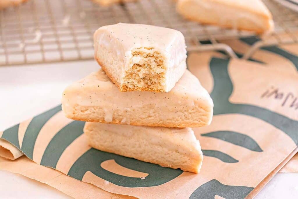 Three vanilla-glazed scones are stacked on a paper bag; the top scone has a bite taken out of it. A cooling rack with more easy desserts is in the background.