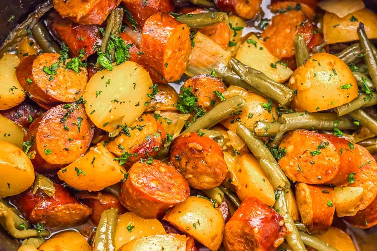 Close-up of cooked sausage slices, potatoes, and green beans mixed with herbs.