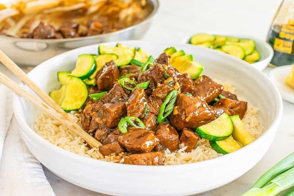 A bowl of rice topped with glazed beef chunks, sliced zucchini, and chopped green onions, with chopsticks on the side—perfect for those craving Simple 30 Minute Meals.