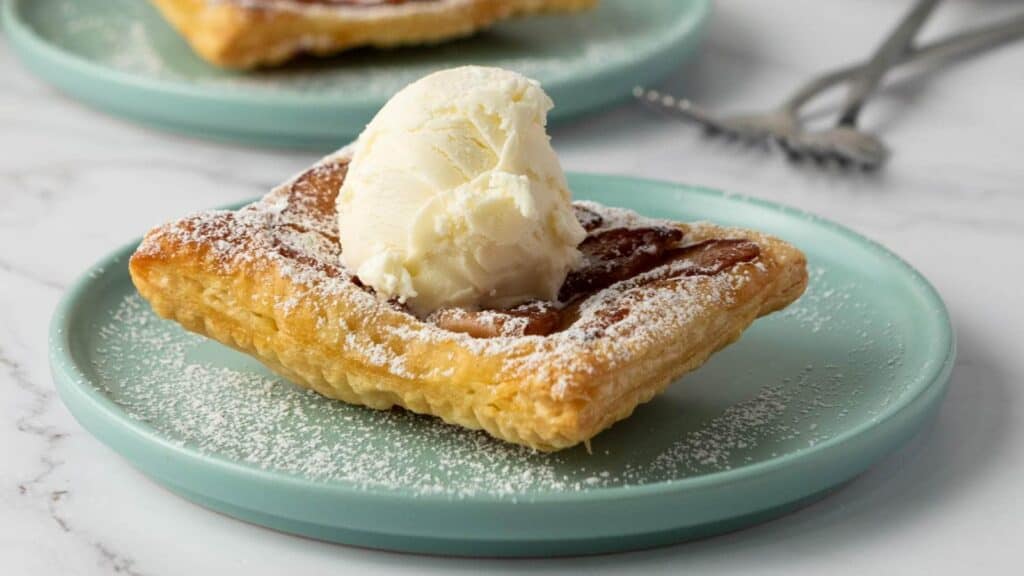 A square piece of puff pastry topped with a scoop of vanilla ice cream and dusted with powdered sugar on a light blue plate.