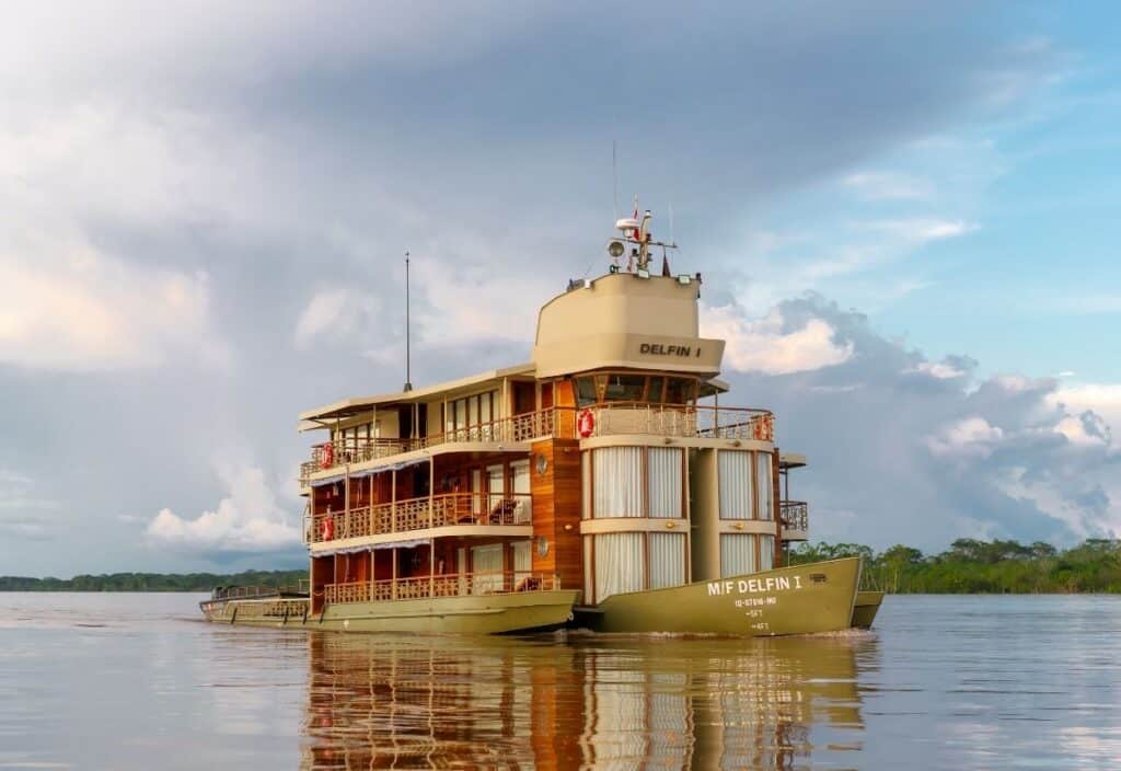 A three-story riverboat named "Delfin II" highlights river cruise growth as it glides on calm water near a forested shoreline under a cloudy sky.