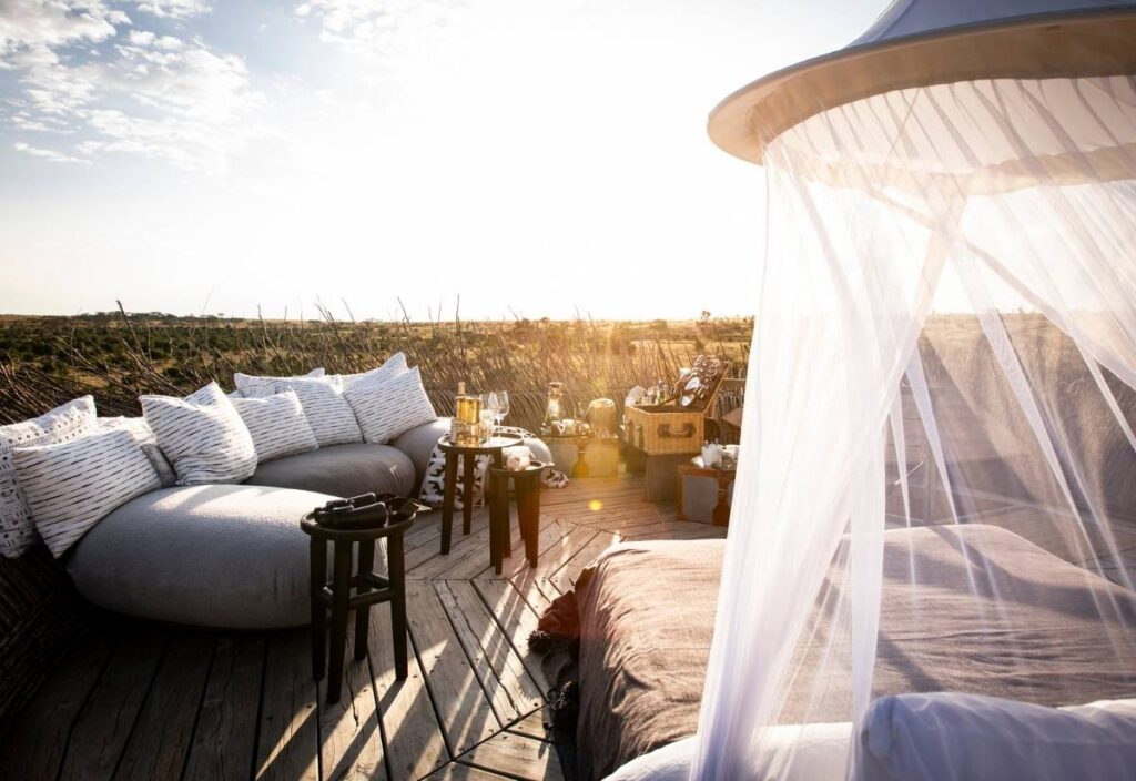 Outdoor lounge area with cushioned seating, a canopy bed with white netting, small tables, and refreshments on a wooden deck surrounded by grass at sunset.