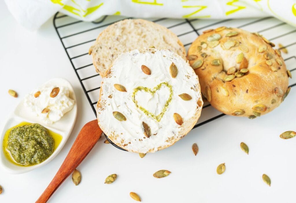 A sliced seed-topped bagel with cream cheese and a pesto heart sits on a cooling rack, alongside a small dish of pesto, olive oil, and cheese. Pumpkin seeds are scattered around.