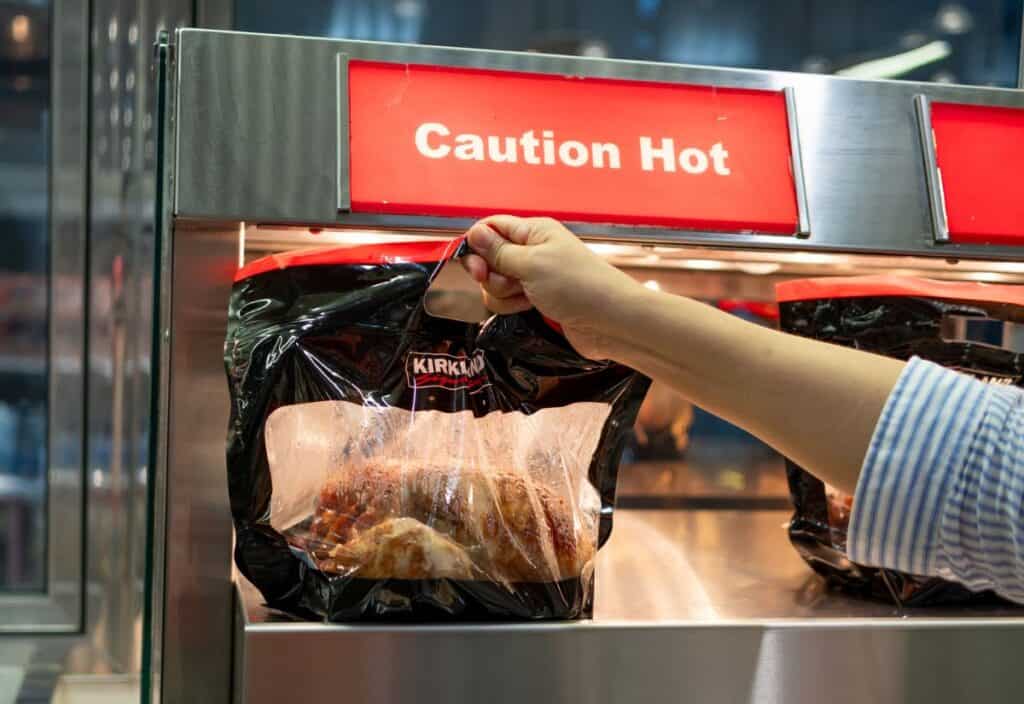 A person picks up a bag of Kirkland rotisserie chicken from a heated display case with a "Caution Hot" sign above it.