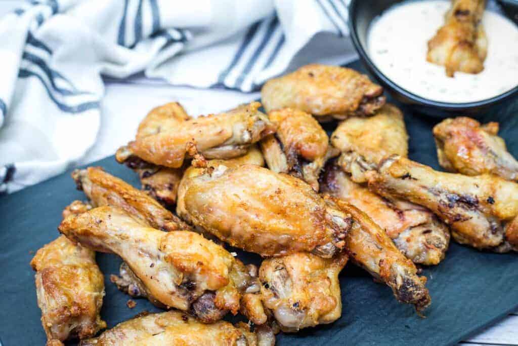 A pile of air-fried chicken wings on a black slate platter with a bowl of creamy dipping sauce in the background.