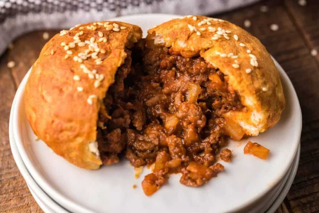 A baked bun with a sesame seed topping is split open, revealing a filling of seasoned ground meat and diced vegetables on a white plate.