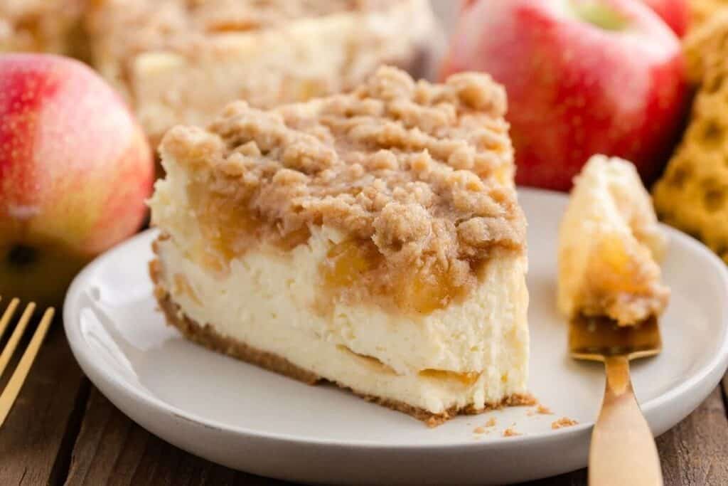 A slice of crumb-topped simple cheesecake with apple filling sits on a plate, surrounded by fresh apples and a gold fork holding a bite of the creamy treat.