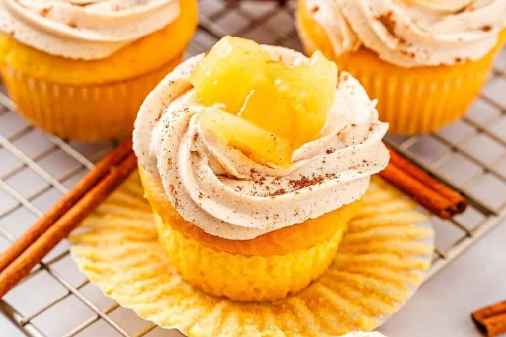 A yellow cupcake with swirled frosting, sprinkled with cinnamon, and topped with pineapple pieces sits on an open cupcake liner next to cinnamon sticks on a cooling rack.