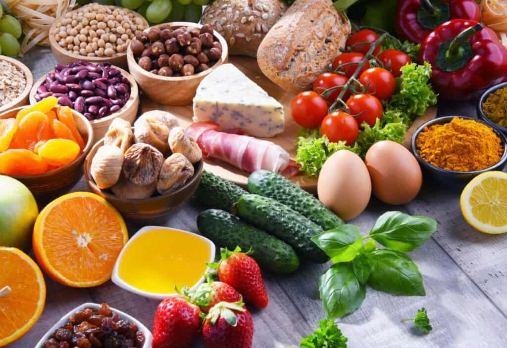 An assortment of fresh fruits, vegetables, eggs, nuts, cheese, bread, dried fruits, herbs, and spices arranged on a wooden surface.