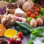 An assortment of fresh fruits, vegetables, eggs, nuts, cheese, bread, dried fruits, herbs, and spices arranged on a wooden surface.