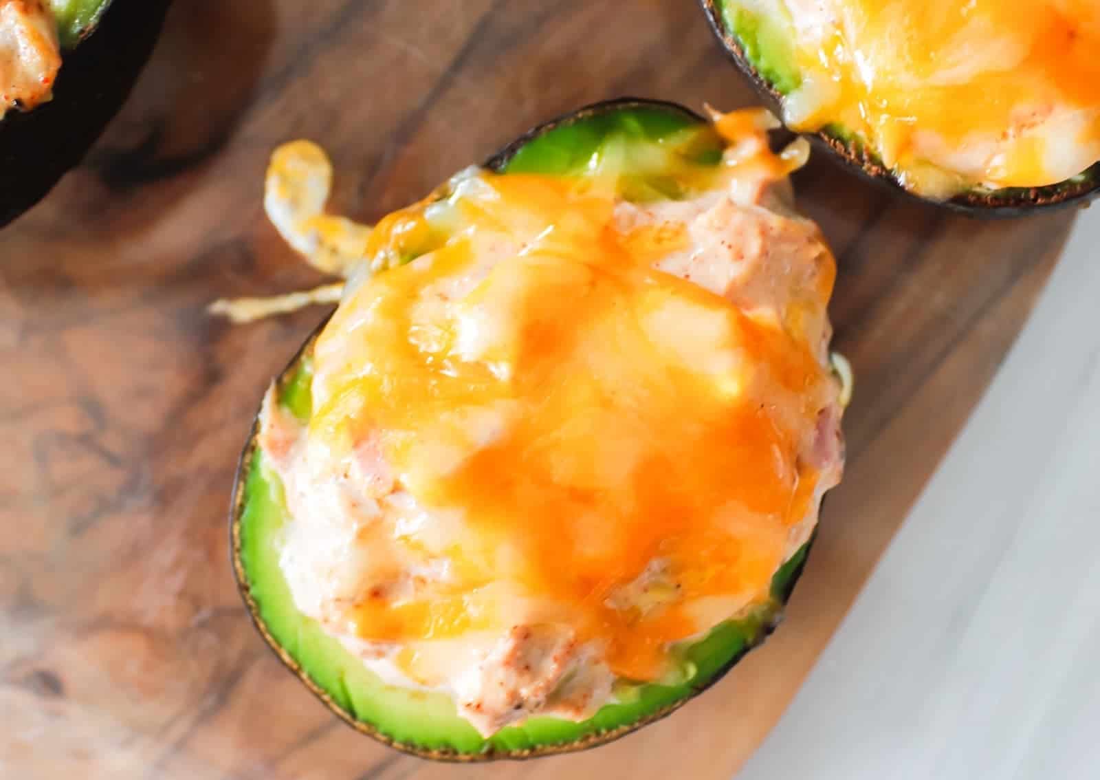 Halved avocado filled with a creamy mixture and topped with melted cheddar cheese, placed on a wooden surface.