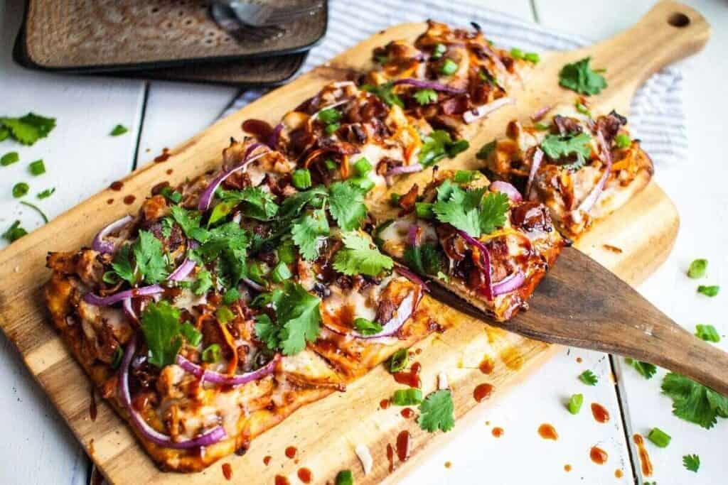 Rectangular flatbread pizza topped with cheese, red onions, green onions, cilantro, and barbecue sauce, presented on a wooden cutting board with a slice being served.
