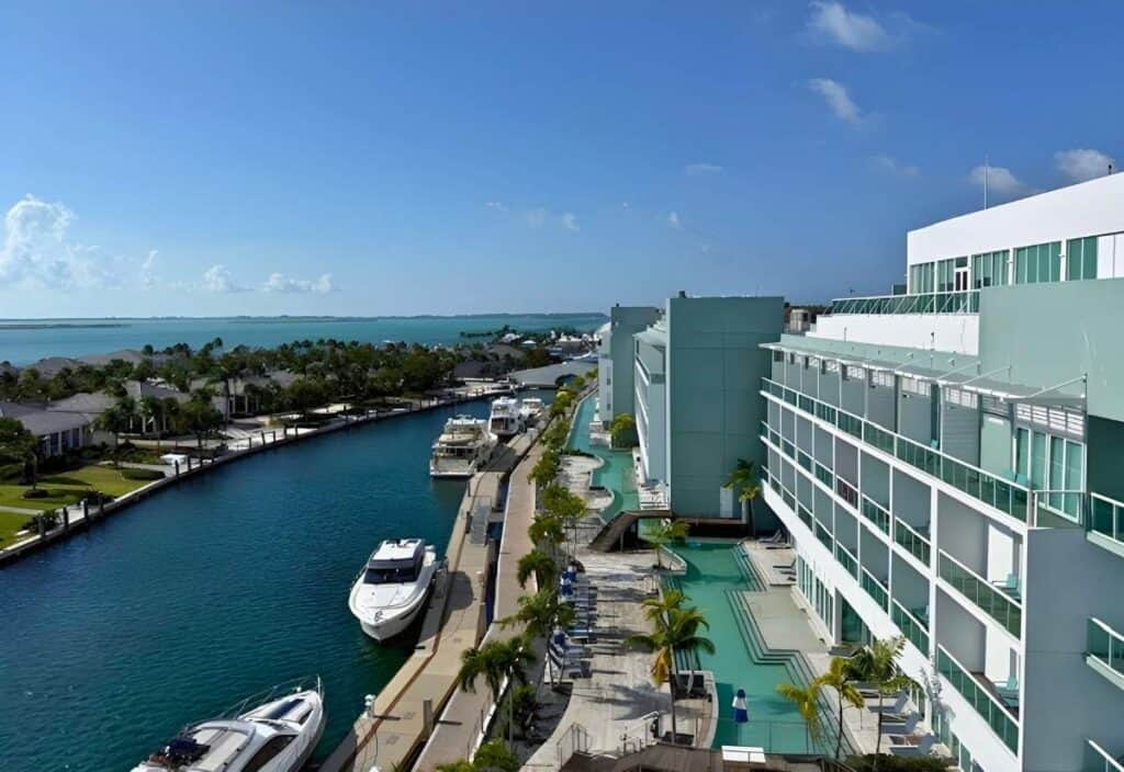 A modern waterfront hotel with balconies overlooks a canal lined with yachts, palm trees, and a pool area; inspired by Bimini, it offers stunning ocean views and blue skies in the background.