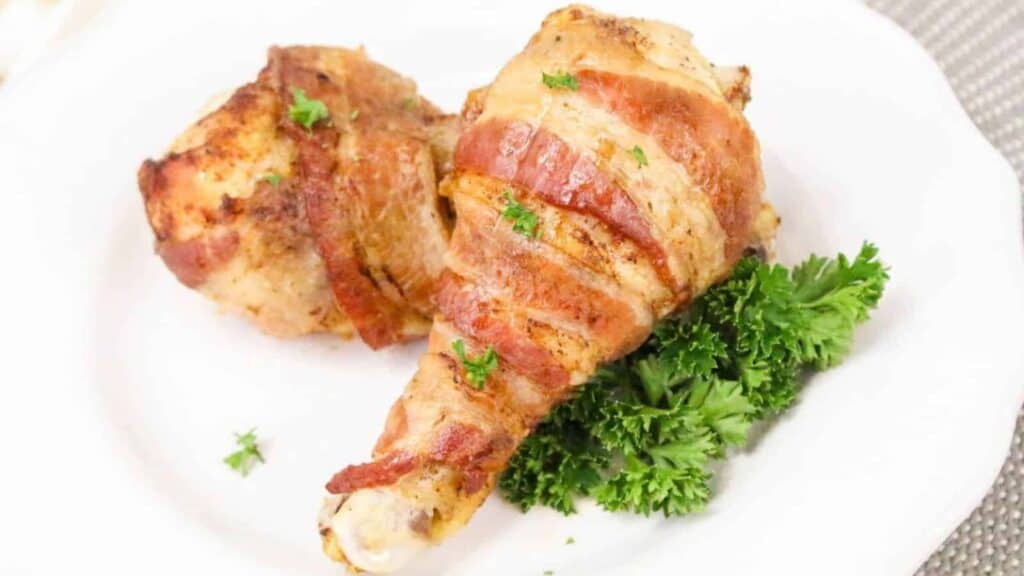 Two baked chicken drumsticks wrapped in bacon are served on a white plate with a garnish of fresh parsley.