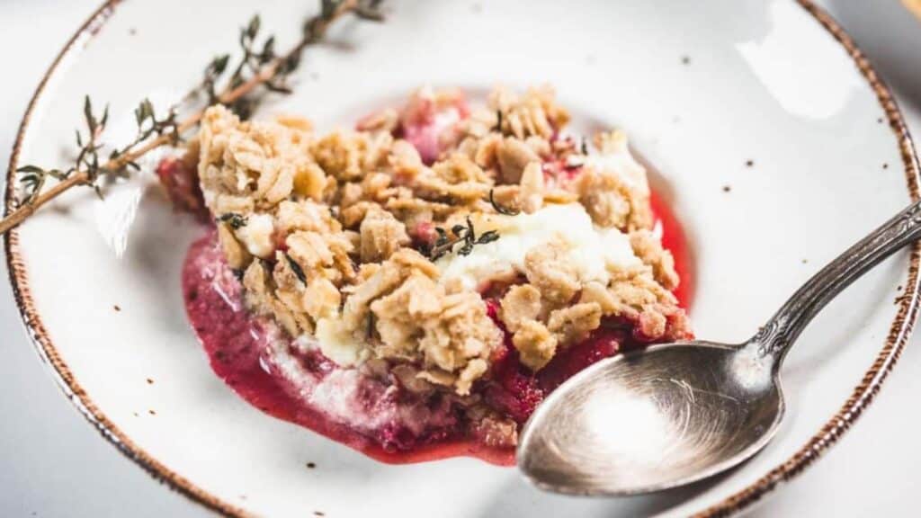 A serving of fruit crumble with oat topping and melted cream sits on a white plate with a silver spoon and a sprig of thyme.