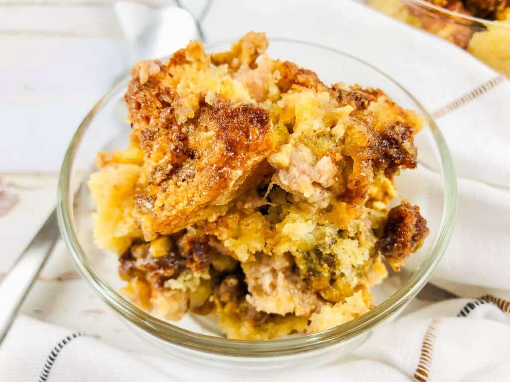 A clear glass bowl filled with a serving of bread pudding, featuring a golden-brown, crumbly texture on top.