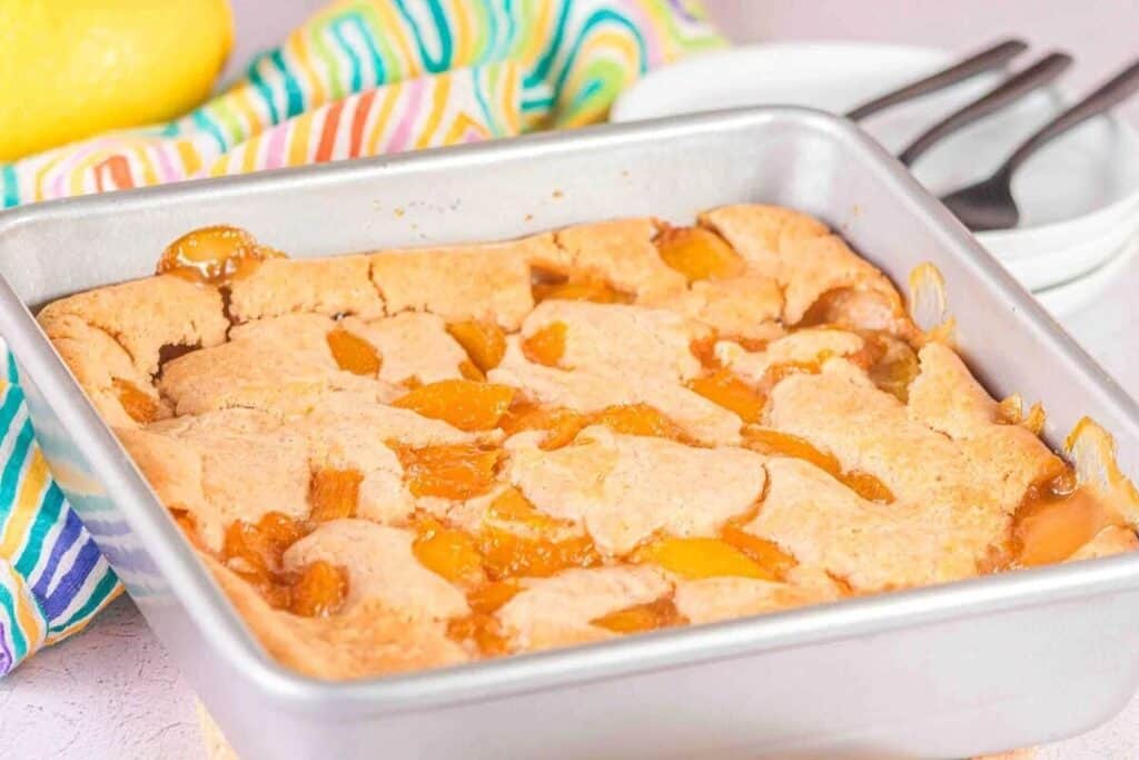 A square metal baking pan filled with peach cobbler sits on a countertop beside stacked white plates, forks, and a striped cloth.