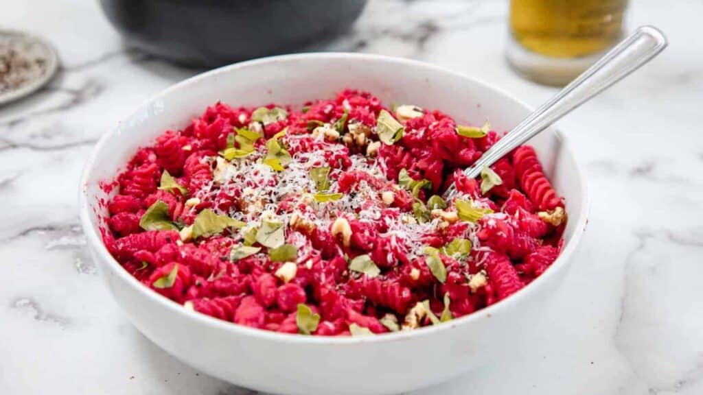 A white bowl filled with pink beet pasta, topped with grated cheese, chopped nuts, and fresh herbs, with a fork resting inside.