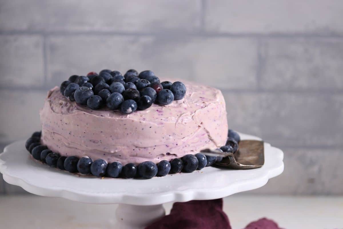 A round cake with blueberry frosting, topped and surrounded by fresh blueberries, sits on a white cake stand with a serving spatula beside it.
