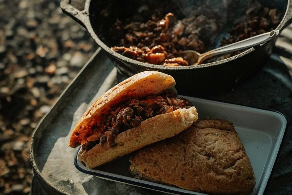 A sandwich filled with saucy meat sits on a tray next to another piece of bread, with a pot of cooked meat and a spoon in the background&mdash;perfect inspiration for your next tasty beef recipes.