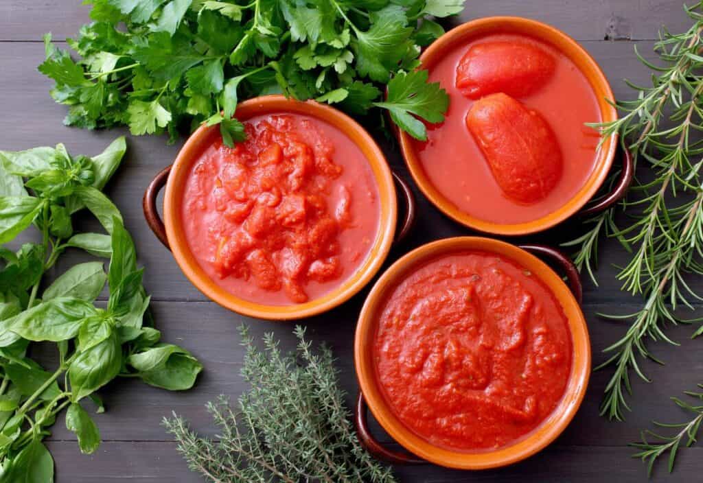 Three bowls of tomato sauce and whole peeled tomatoes are arranged on a wooden surface, surrounded by fresh basil, parsley, rosemary, and thyme.