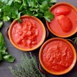 Three bowls of tomato sauce and whole peeled tomatoes are arranged on a wooden surface, surrounded by fresh basil, parsley, rosemary, and thyme.